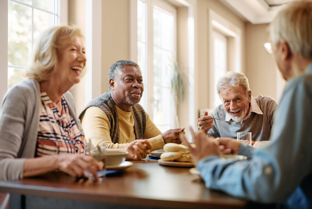 Happy African American senior man talking to his friends while eating at dining table at nursing home.